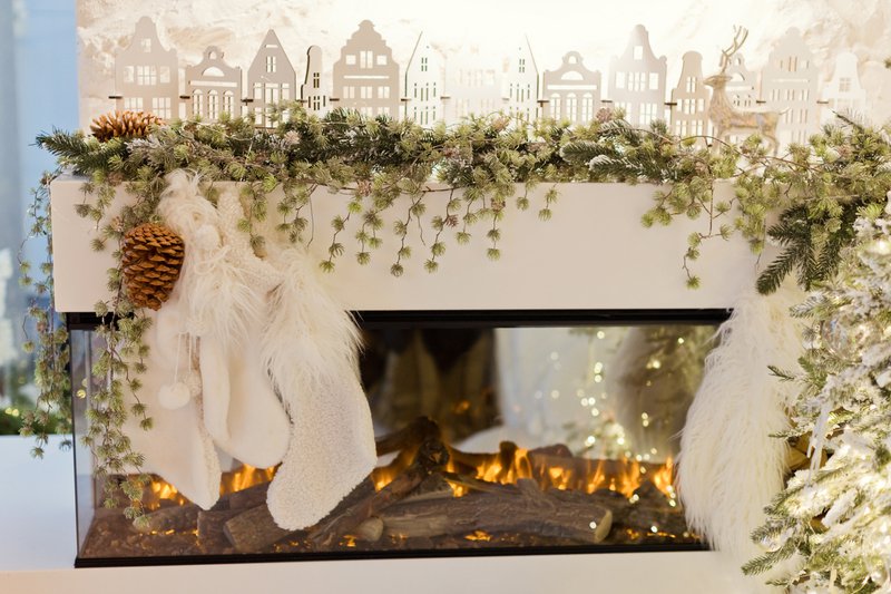 White interior with modern fireplace with burning logs, decorated Christmas garland, socks and houses near Christmas tree