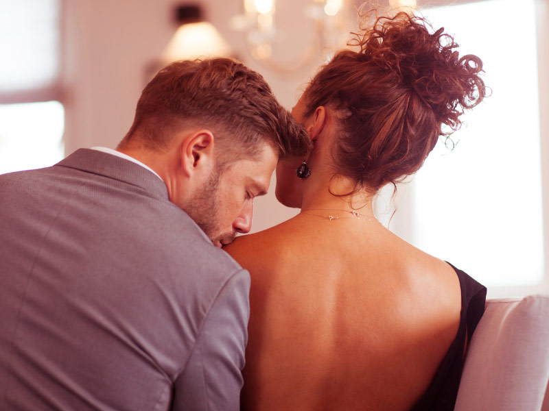 a photo of a couple in evening wear sitting together, and the man is kissing the woman's shoulder